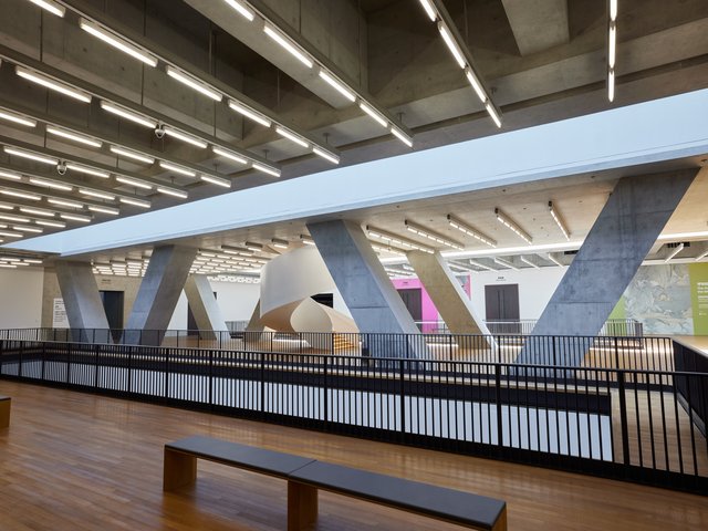 View of a large space with wooden floors. Diagonal concrete pillars in a truss structure line the centre of the space on either side of a spiral staircase. Railings outline an opening in the floor in front of us.